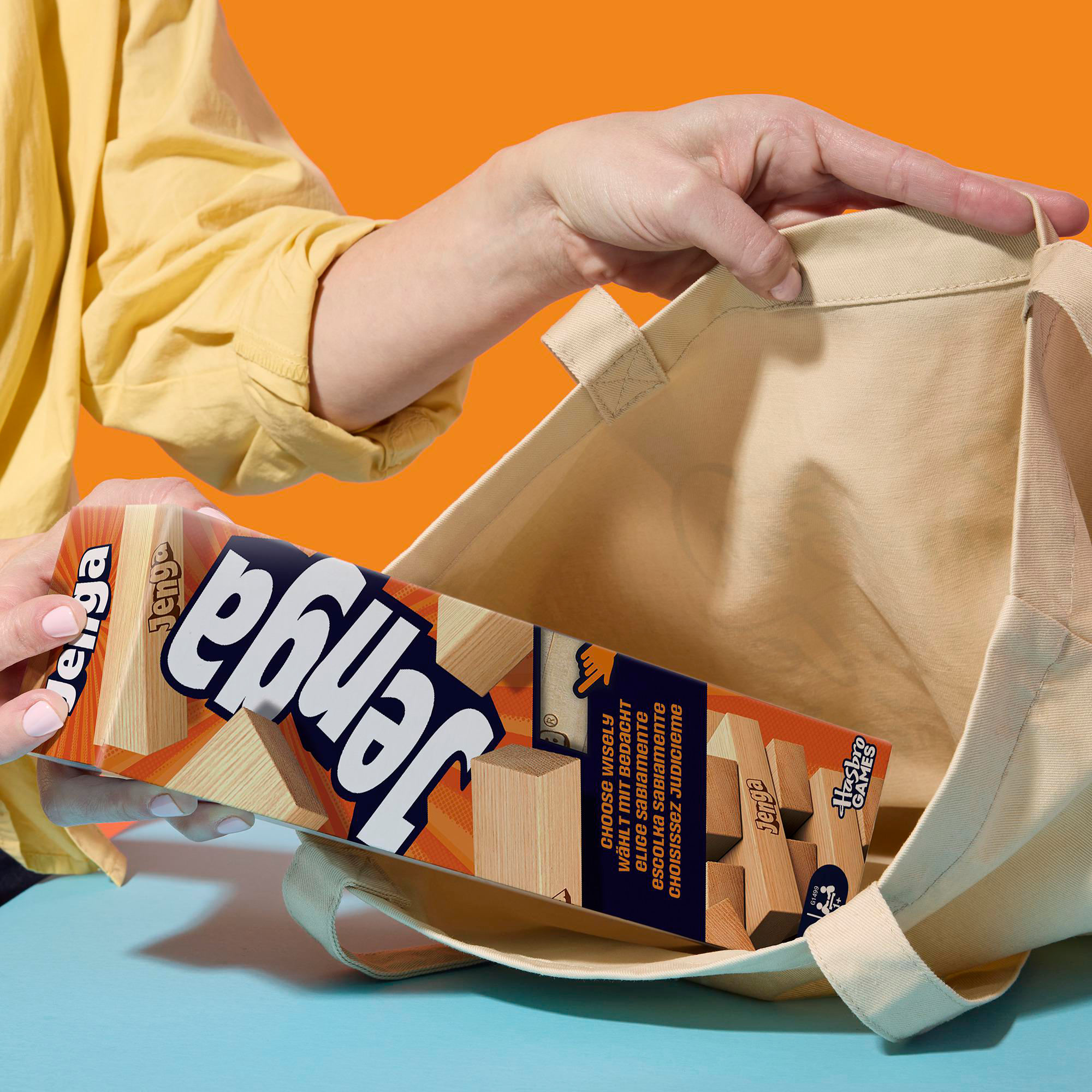 Eine Person hält eine Jenga-Spielbox und legt sie in eine beigefarbene Einkaufstasche. Der Hintergrund ist orange, mit blauer Oberfläche.