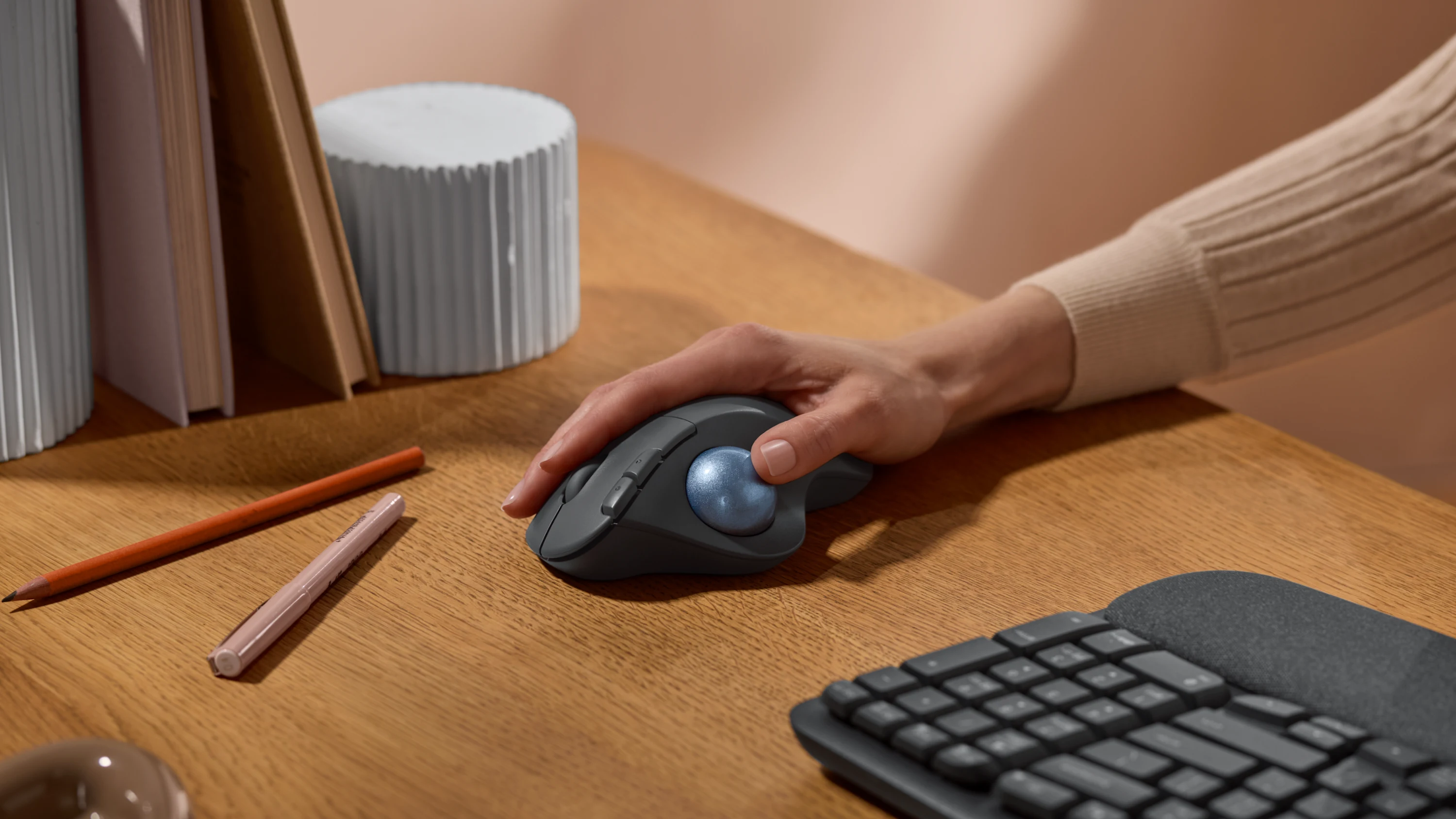 Une main utilise une souris trackball sur un bureau en bois. Il y a aussi des livres, des crayons et un clavier. Le fond est uni.