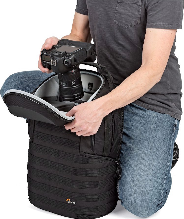 Una persona è in ginocchio e mette una fotocamera in uno zaino nero. La persona indossa jeans e una maglietta grigia. Lo zaino ha un logo Lowepro.
