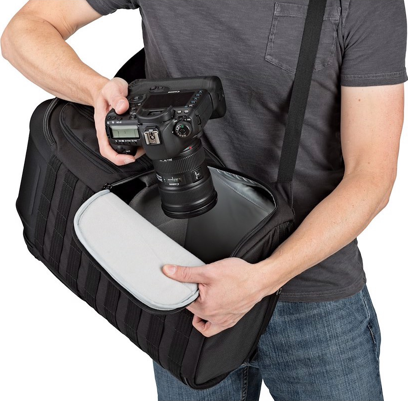 Una persona tiene in mano una fotocamera nera e una borsa per fotocamera. La borsa è aperta, rivelando un'imbottitura. La persona indossa una maglietta grigia e jeans.