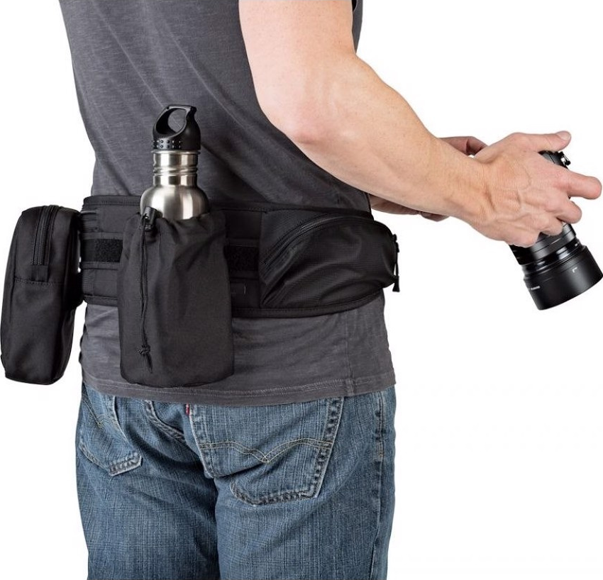 Una persona indossa una cintura multiuso nera con tasche e una bottiglia d'acqua. Tengono una fotocamera nella mano destra, con i jeans.
