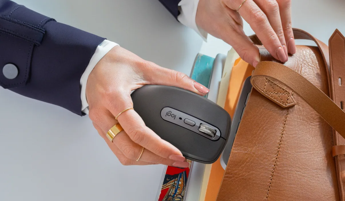 Des mains tiennent une souris grise à côté d'un sac en cuir marron et de cahiers colorés.