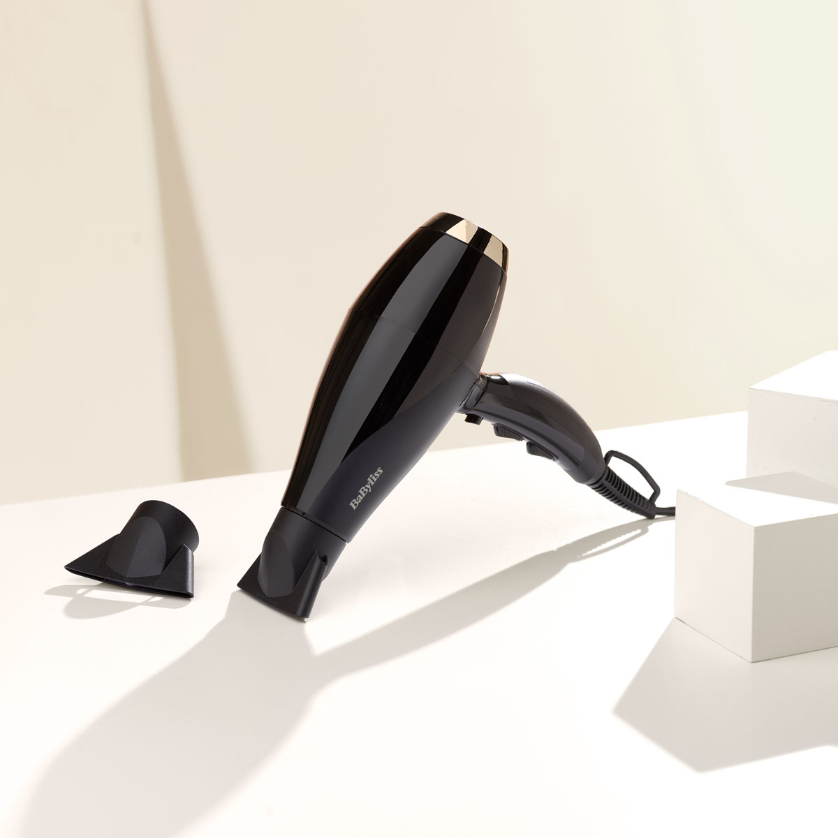 Ein schwarzer Haartrockner mit Düse auf einer weißen Oberfläche. Weißer Hintergrund und Schatten schaffen eine minimalistische Ästhetik.