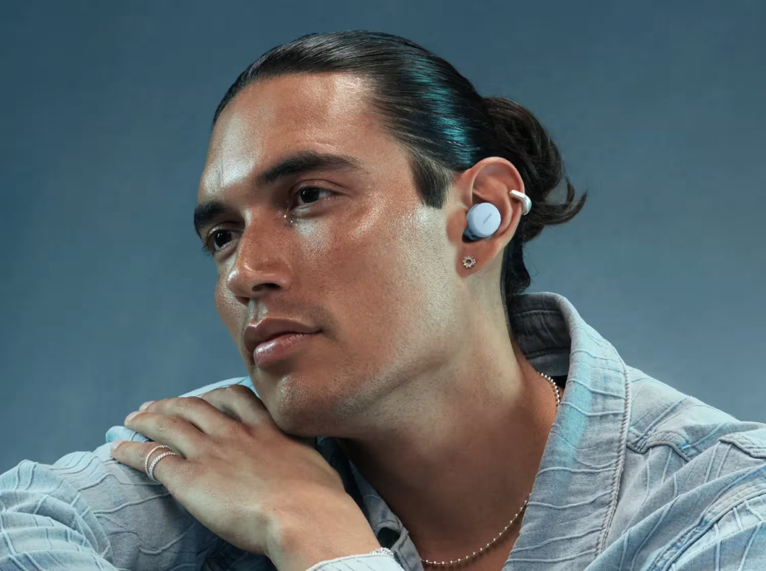 Una persona con capelli e pelle scura indossa auricolari blu chiaro. Hanno una collana a catena e una camicia di jeans.
