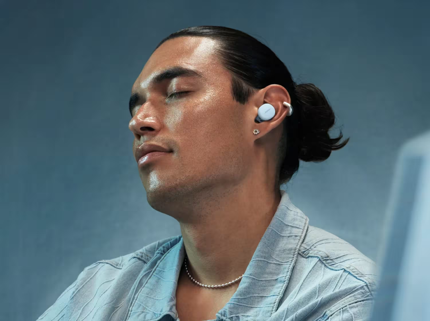 Un uomo con gli occhi chiusi indossa auricolari, una catena d'argento e una camicia. I suoi capelli sono legati. Lo sfondo è un gradiente blu.