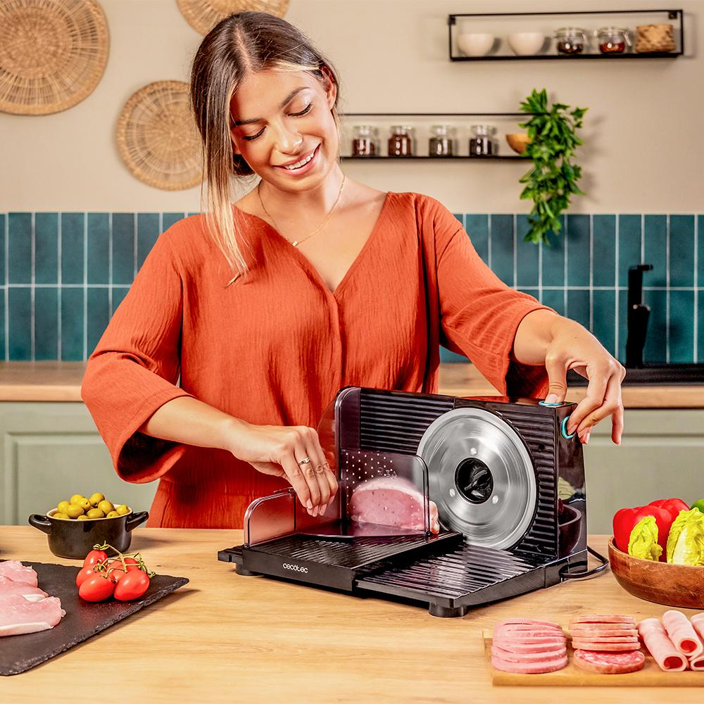 Una mujer corta carne con una cortadora. Varios ingredientes están sobre la mesa, como tomates, aceitunas y lechuga.