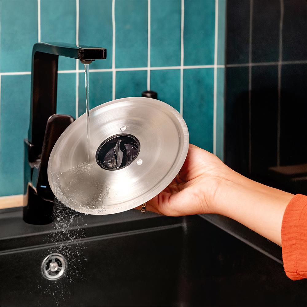 Una mano sostiene una herramienta de cocina de metal bajo agua corriente de un grifo negro, sobre un fregadero negro. Azulejos turquesa en el fondo.