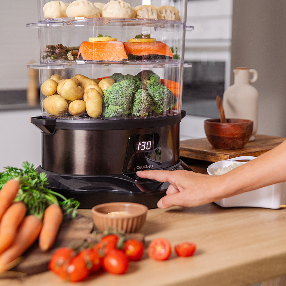 Una vaporera de varios niveles con comida dentro. Se ven salmón, verduras y albóndigas. Una mano pulsa un botón. Zanahorias y tomates en primer plano.