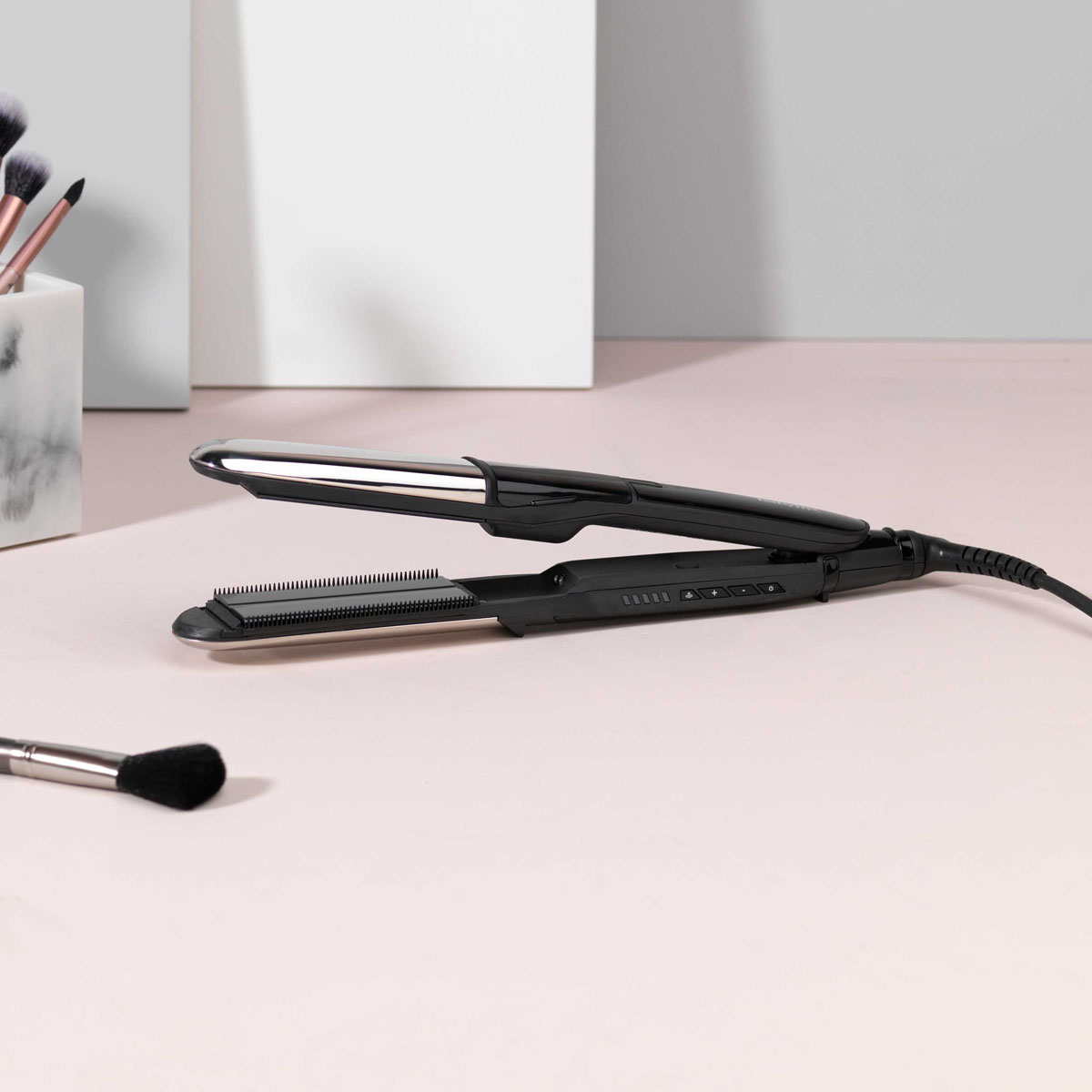 Schwarzes Glätteisen auf rosa Oberfläche. Make-up-Pinsel im Hintergrund, ein Pinsel im Vordergrund. Weißer Hintergrund.