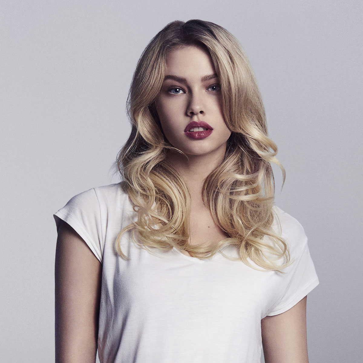 Blonde Frau in weißem T-Shirt, roter Lippenstift, blickt direkt in die Kamera. Weicher Hintergrund.