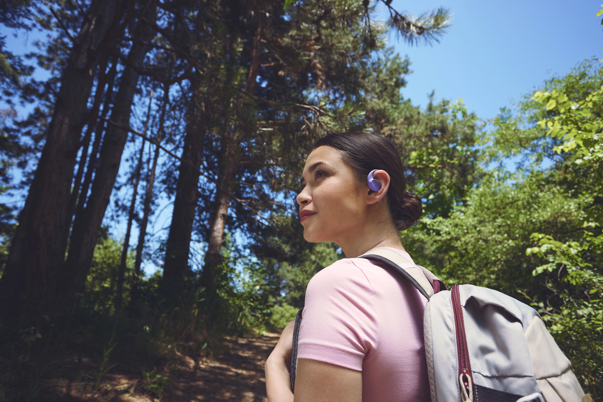 Eine Frau mit lila Ohrhörern und Rucksack wandert in einem Wald. Bäume sind im Hintergrund, der Himmel ist blau.