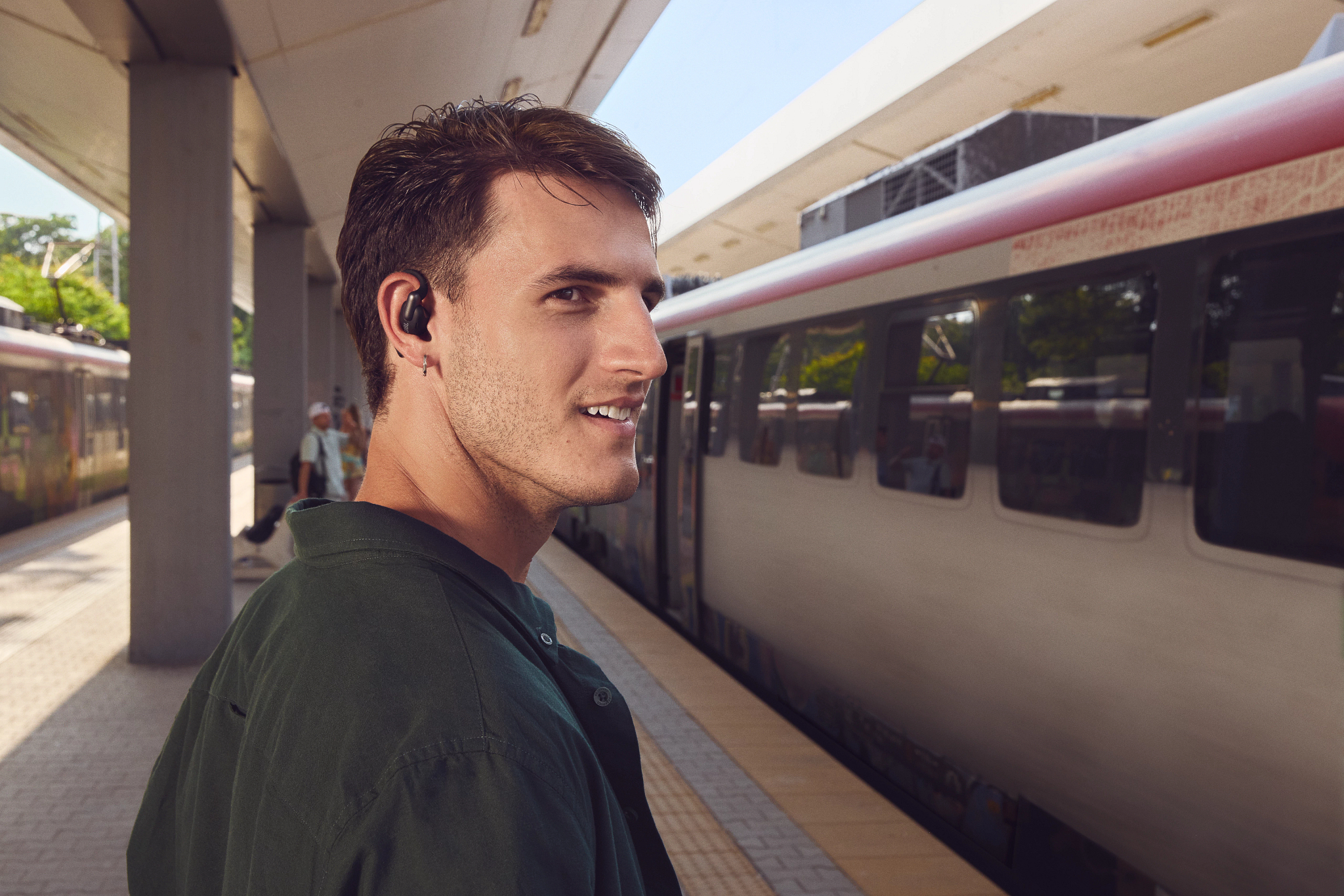 Ein Mann mit Ohrhörer steht lächelnd auf einem Bahnsteig. Ein Zug steht hinter ihm, ein weiterer ist im Hintergrund zu sehen.