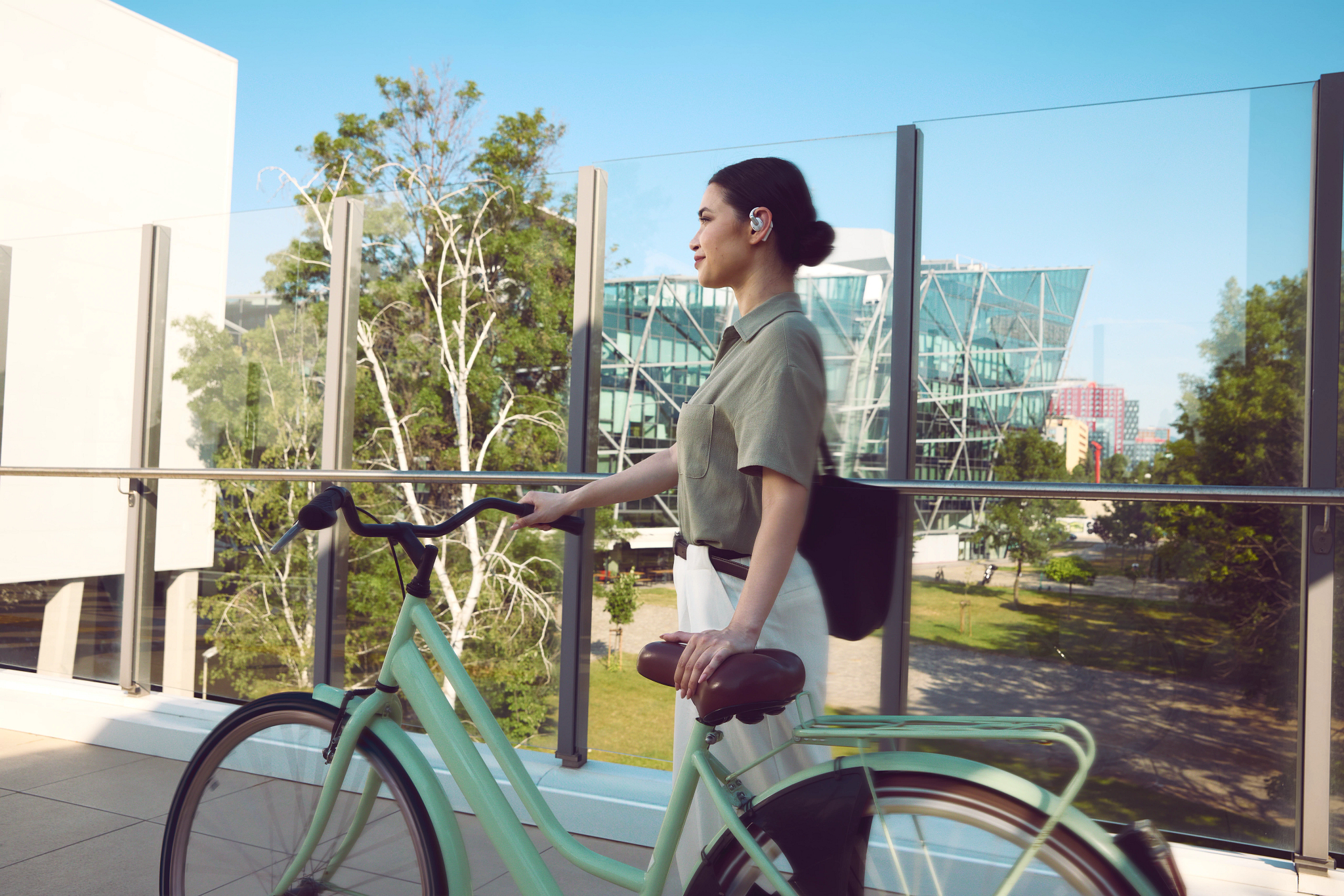 Eine Frau geht mit einem hellgrünen Fahrrad. Sie trägt ein grünes Hemd, eine weiße Hose und hat eine schwarze Tasche. Gebäude und Bäume sind im Hintergrund.