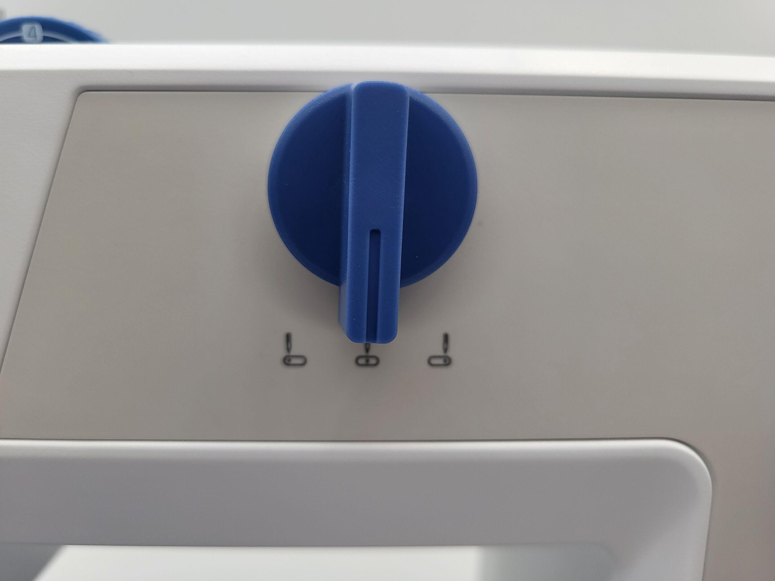 Nahaufnahme zeigt einen blauen Knopf auf weißer Oberfläche. Darunter kleine schwarze Symbole.