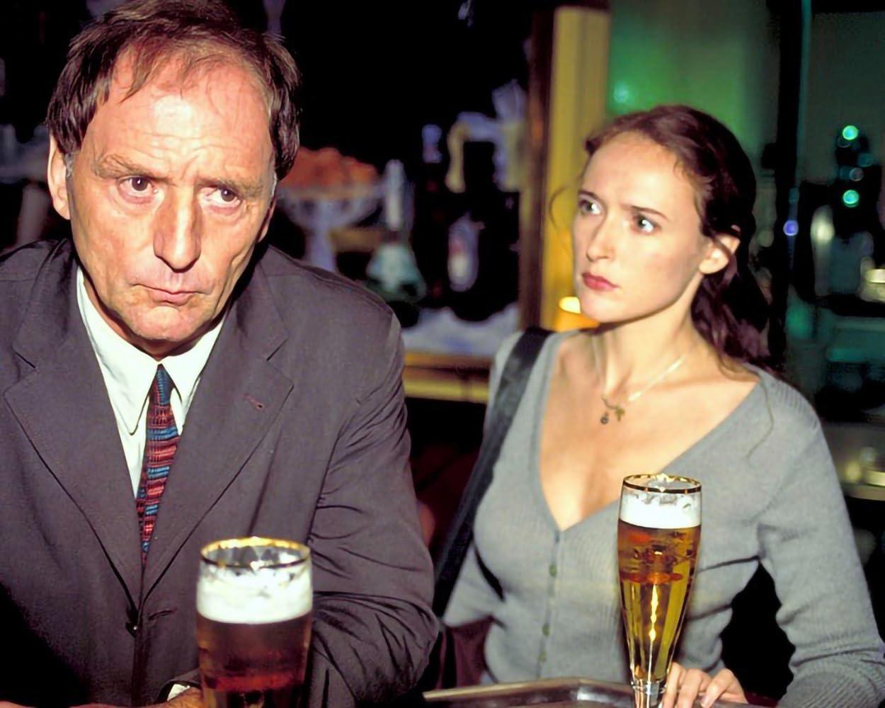 Ein Mann im Anzug und eine Frau in einem grauen Pullover in einer Bar. Beide schauen in die Kamera. Zwei Biere auf dem Tisch.