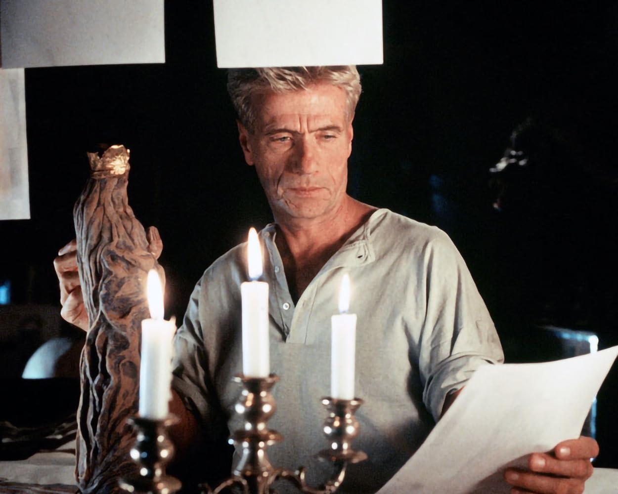 Ein Mann hält ein Papier und eine Holzstatue mit goldener Krone, beleuchtet von Kerzen. Ein dunkler Hintergrund vervollständigt die Szene.