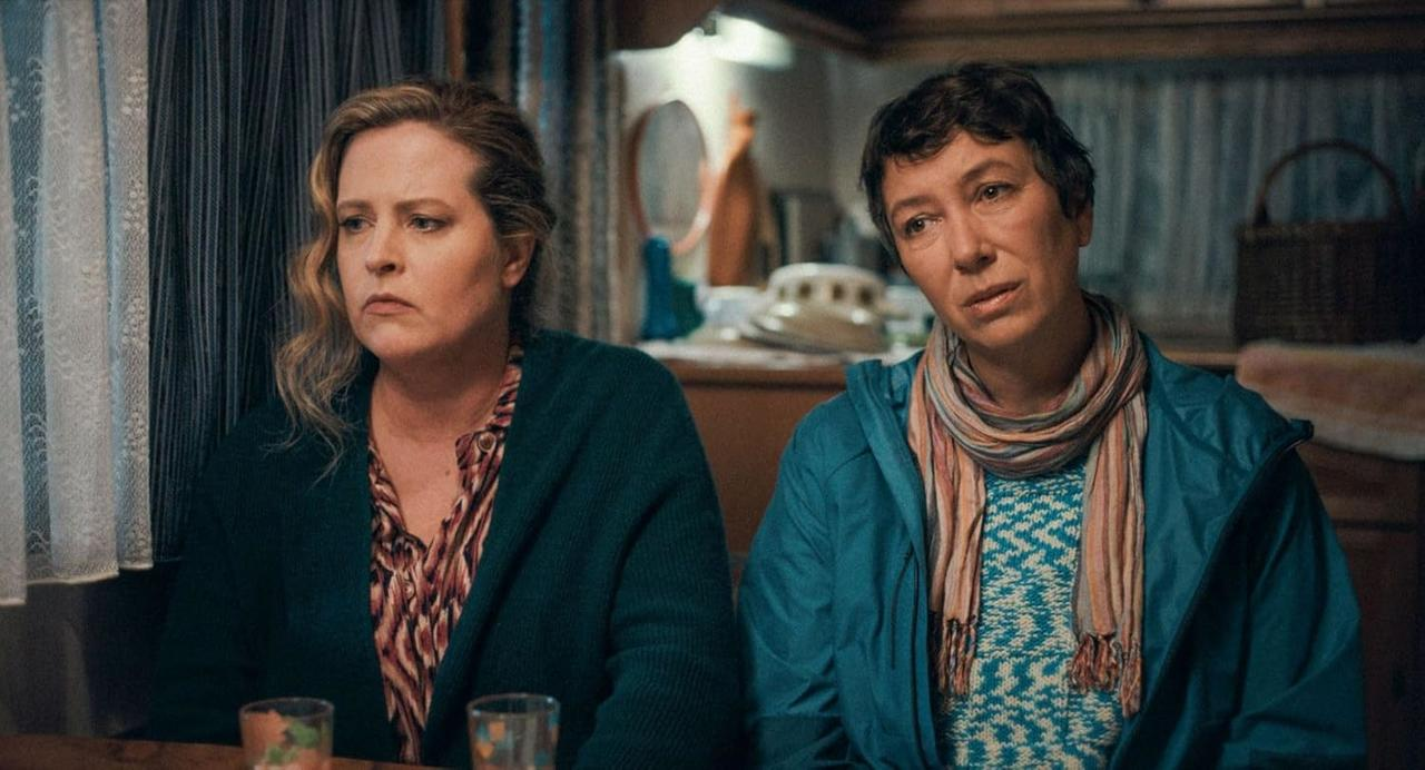 Zwei Frauen sitzen nach vorne. Eine trägt eine grüne Strickjacke und ein gemustertes Hemd. Die andere trägt eine blaue Jacke und einen Schal.