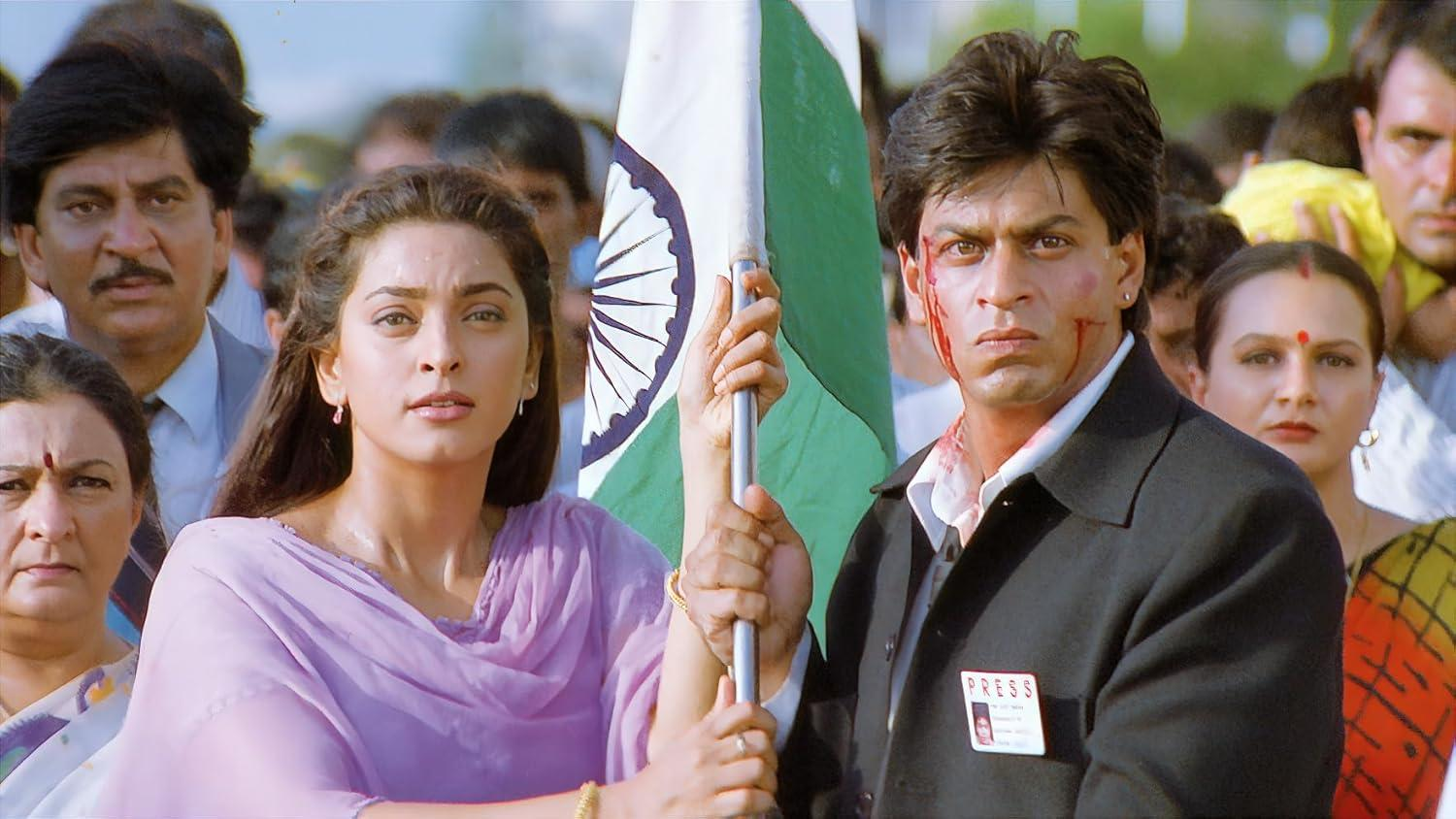 Eine Filmszene zeigt einen Mann und eine Frau, die eine indische Flagge halten. Der Mann hat ein blutiges Gesicht. Viele Menschen sind im Hintergrund.