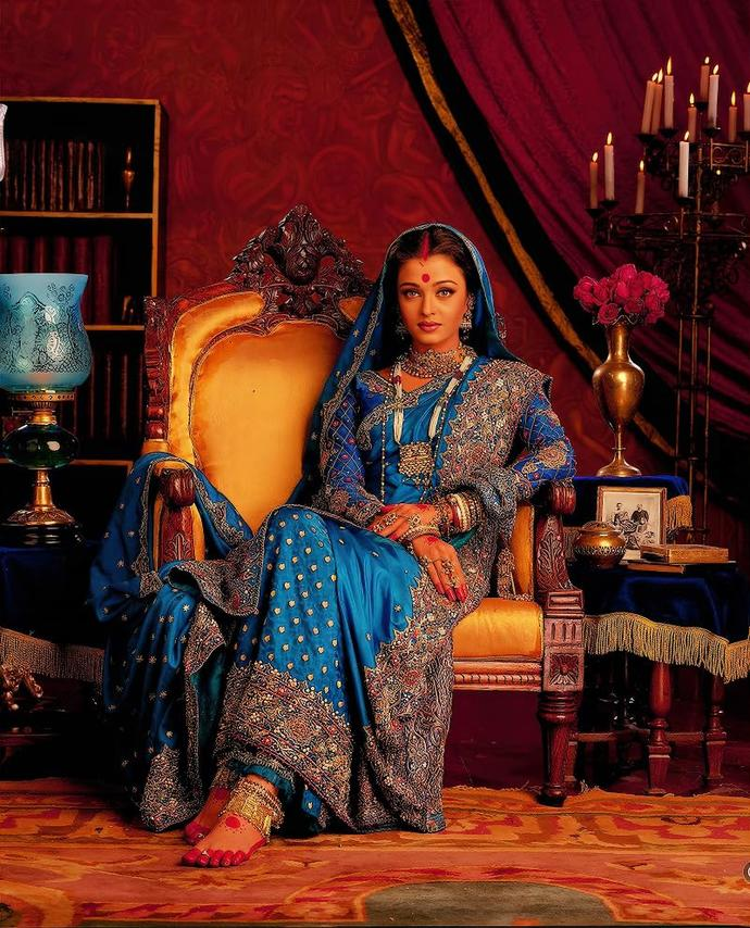 Frau in blauer traditioneller Kleidung sitzt auf einem goldenen Stuhl. Roter Bindi, Schmuck und roter Hintergrund mit Kerzen und Tisch.