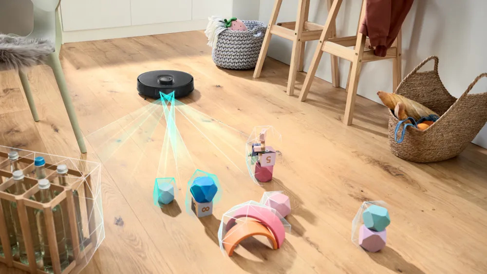 Robot odkurzający porusza się wokół drewnianych klocków na drewnianej podłodze. Widoczny jest również kosz, krzesło i stojak na butelki z wodą.