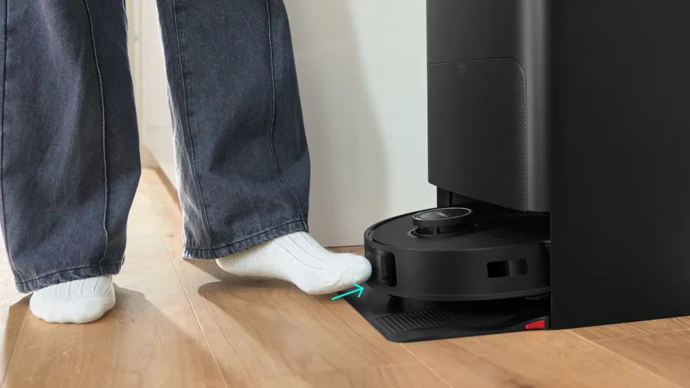 Stopa osoby w białej skarpetce dotyka czarnego robota sprzątającego na drewnianej podłodze. Obecna jest czarna stacja ładująca.