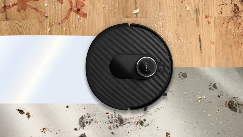 Czarny robot sprzątający na podłodze z plamami i śladami łap. Podłoga podzielona jest na sekcje drewniane i betonowe.