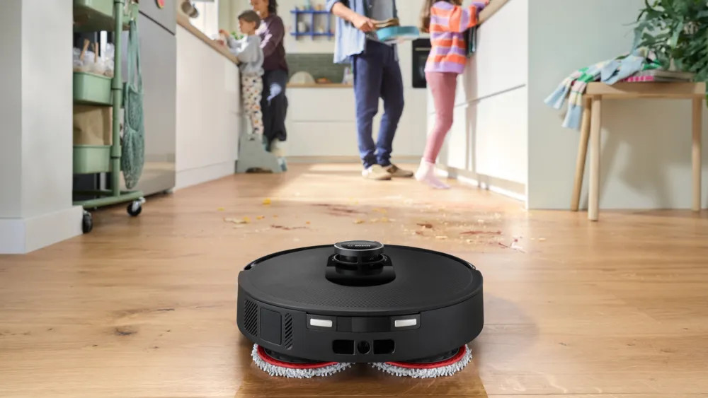 Czarny robot odkurzający sprząta nieuporządkowaną drewnianą podłogę. Ludzie są w tle. Jedzenie i płyny są rozlane na podłodze.