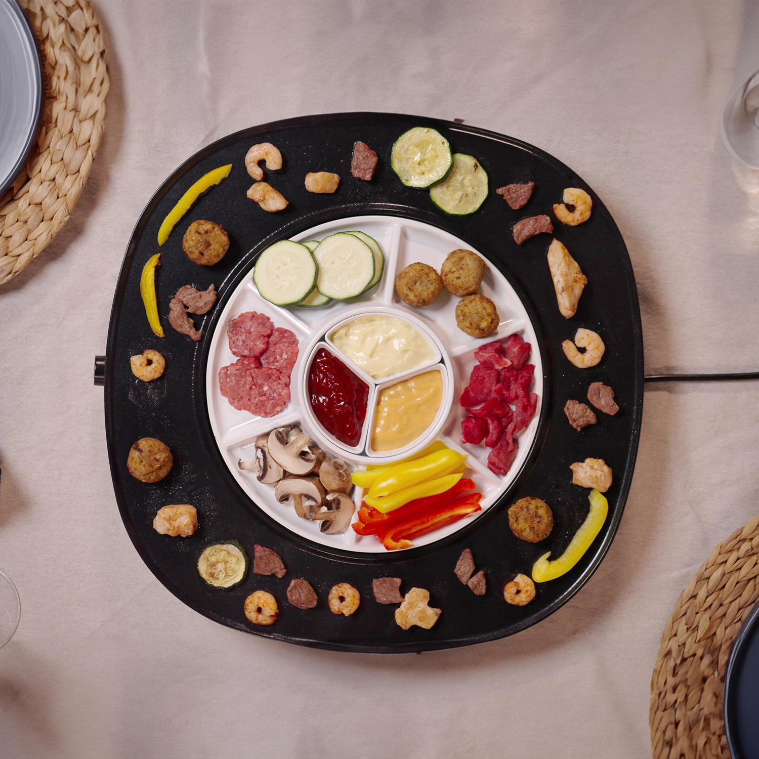 A fondue set with various ingredients: shrimp, meat, vegetables, and sauces arranged around a central white compartment on a black plate.