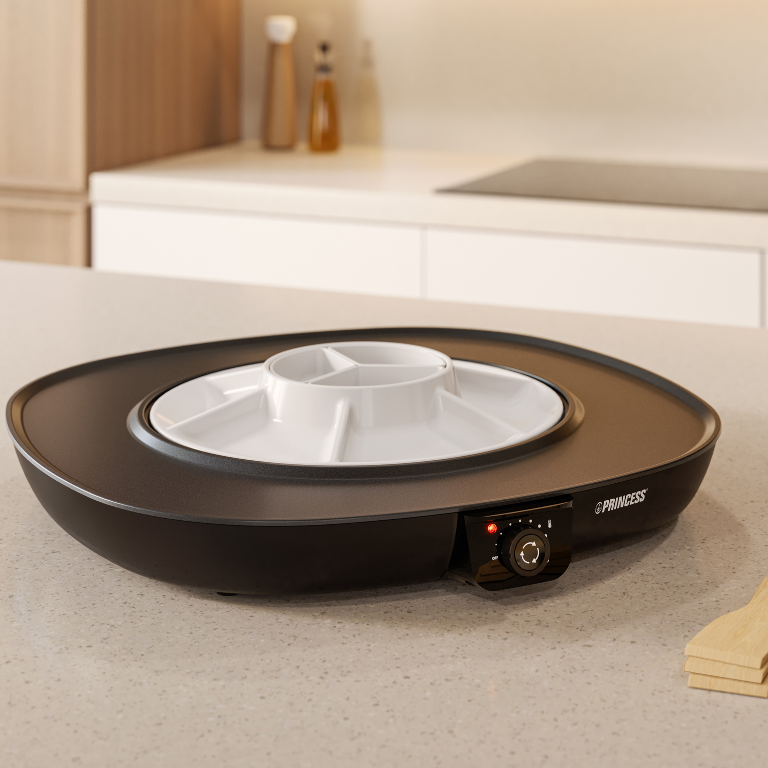 A black Princess fondue set with a white divided serving dish on a light countertop. Kitchen background with cabinets and appliances.