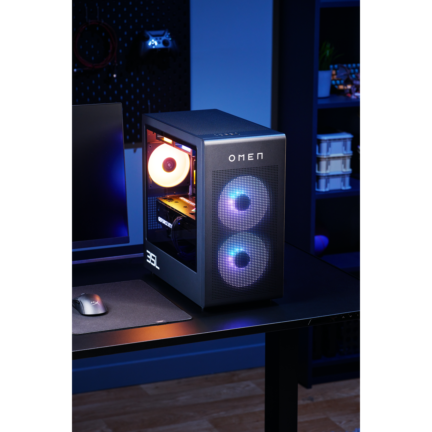 A gaming PC with lit fans, OMEN logo, on a black desk. Monitor and mouse are visible. Dark blue backdrop with shelves in the background.
