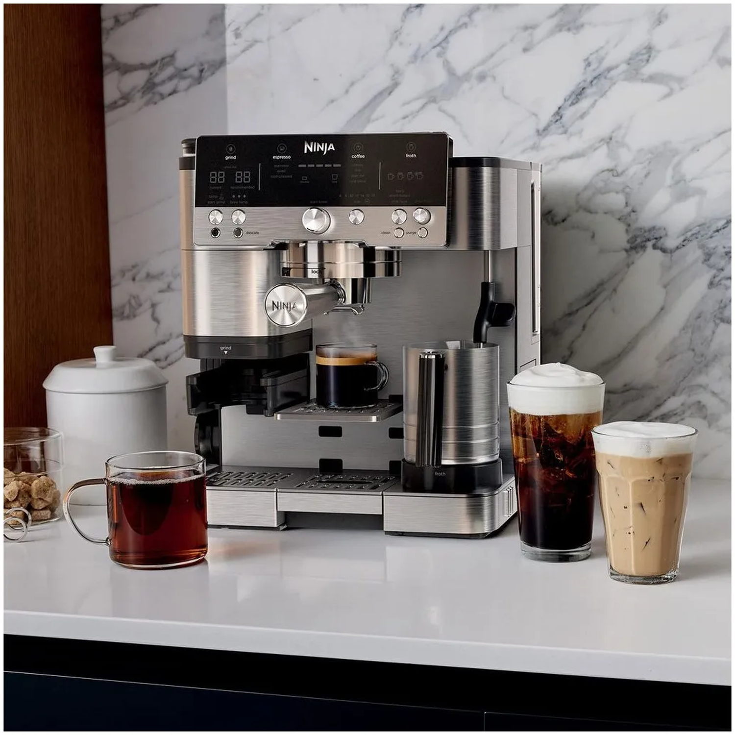 Een Ninja koffiezetapparaat met verschillende koffiedranken en een glazen mok op een wit aanrechtblad tegen een marmeren achterwand.