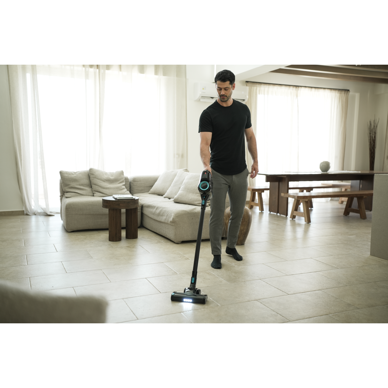 A man vacuums a tiled floor with a black and teal cordless vacuum. A sofa, table, and dining area are in the background.