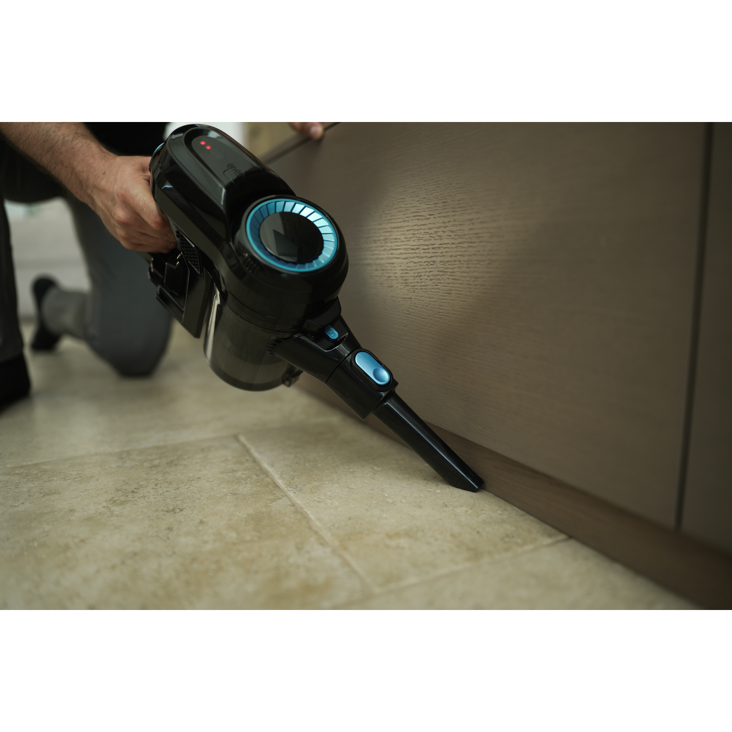 A person is using a black and blue handheld vacuum cleaner. The vacuum is cleaning near a wooden cabinet and beige tile floor.