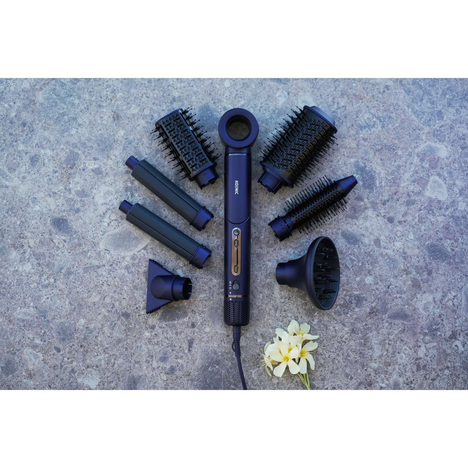A dark blue hair dryer with attachments, arranged on a grey stone surface, with white flowers at the bottom right.