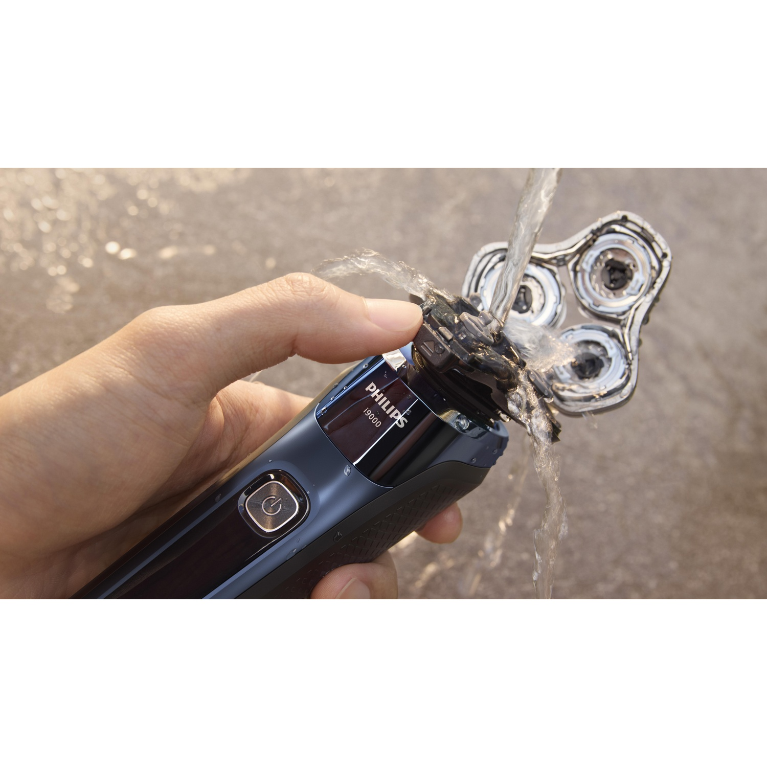 A hand holds a Philips shaver being rinsed with water. Water streams over the shaver head. The background is a blurry surface.