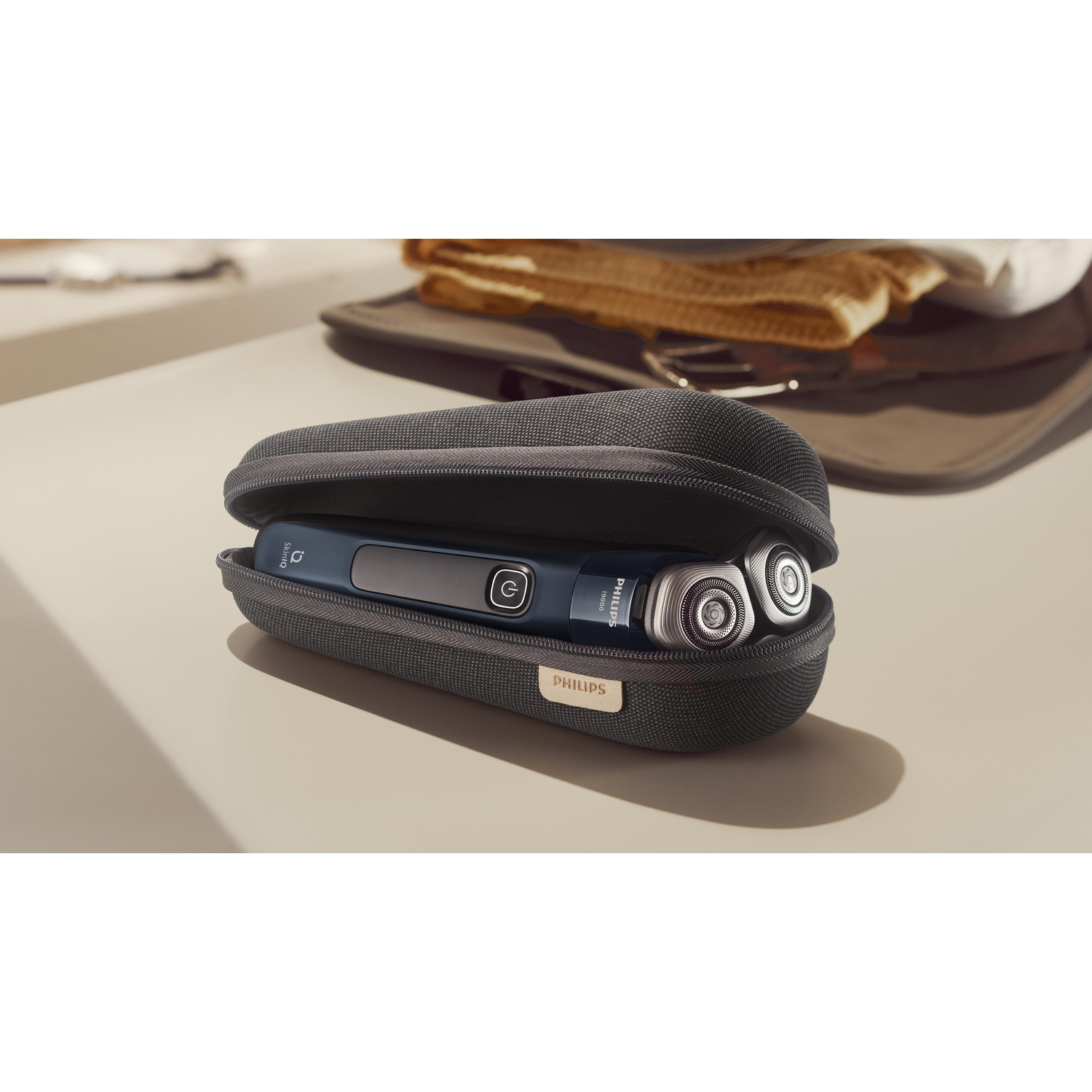 A dark gray travel case contains a blue and silver Philips shaver. The case is open on a light beige surface.
