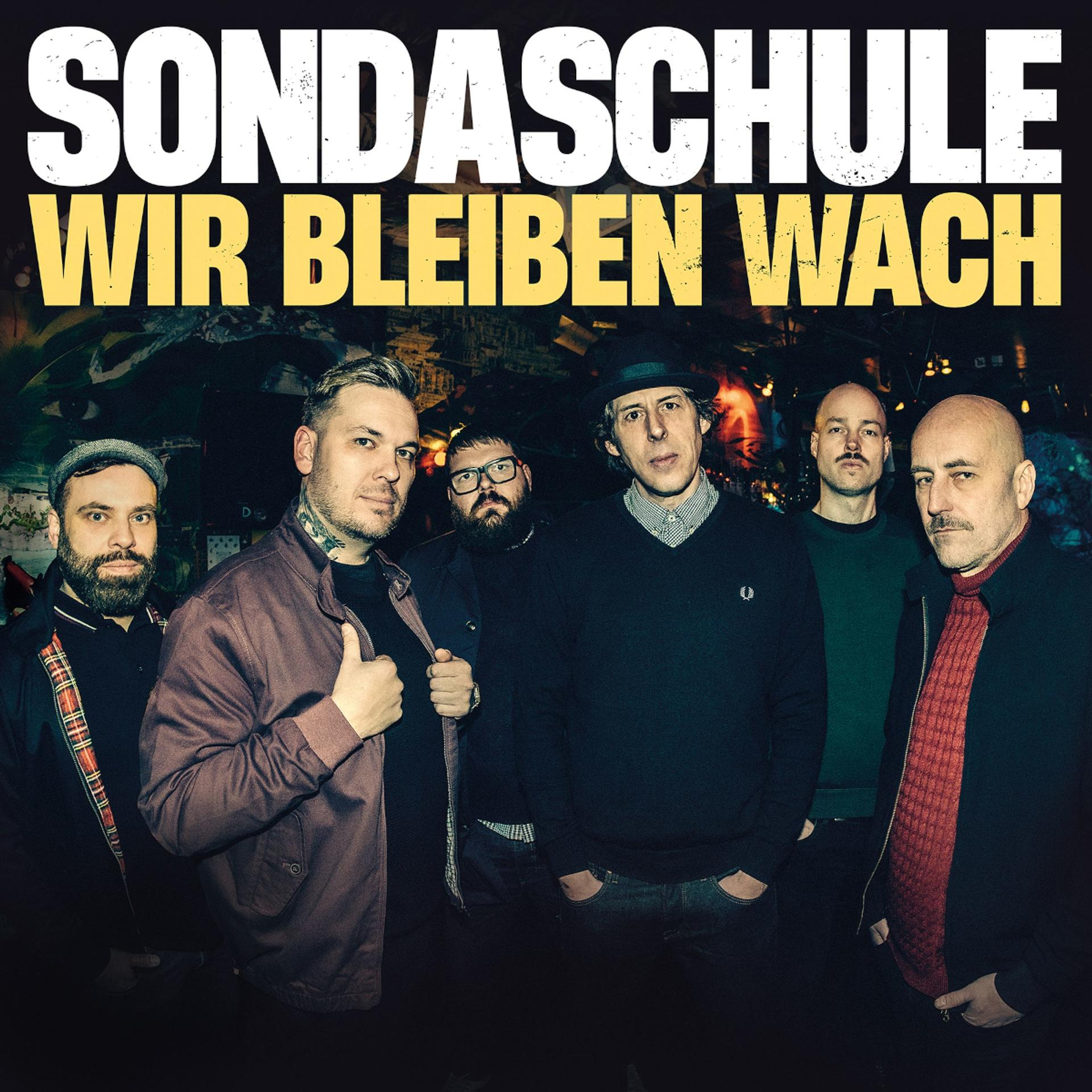 Sechs Männer stehen in einer Gruppe. Die Band Sondaschule. Text lautet: Sondaschule Wir Bleiben Wach.