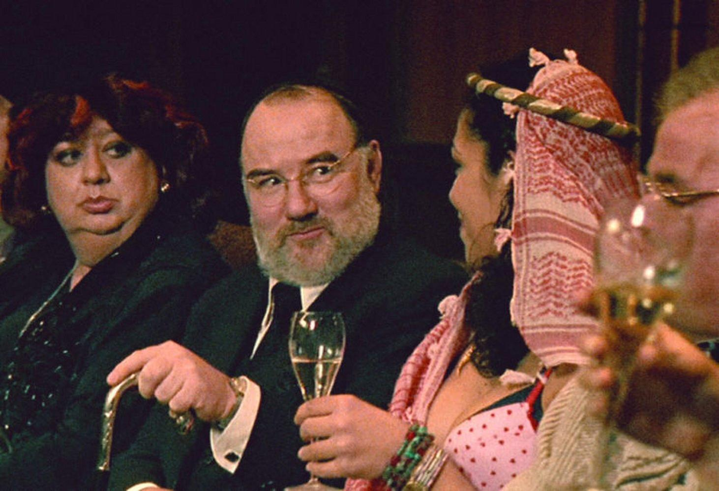 Gruppe von Leuten bei einer Versammlung. Mann mit Brille und Bart hält ein Glas. Frau mit rosa Kopftuch. Andere sind sichtbar.