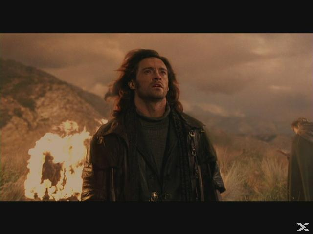 Ein Mann mit langen Haaren blickt nach oben, trägt einen Ledermantel, Feuer links, bewölkter Himmel und Berge im Hintergrund.