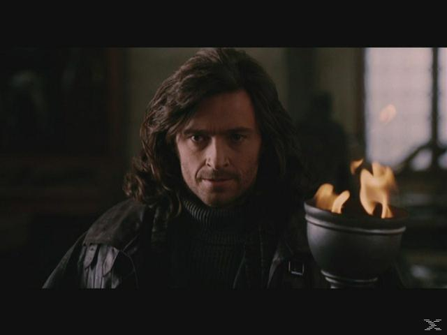 Ein Mann mit langen Haaren hält eine Fackel mit Flammen. Er trägt einen dunklen Pullover und eine Jacke. Der Hintergrund ist schwach beleuchtet.