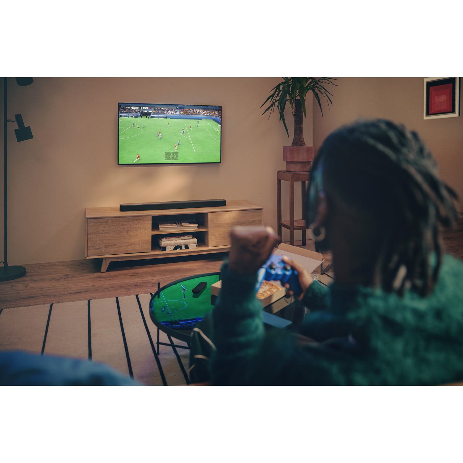 Une personne joue à un jeu vidéo, regardant un match de football à la télé. Une pizza et une manette sont au premier plan. La pièce a un design moderne.