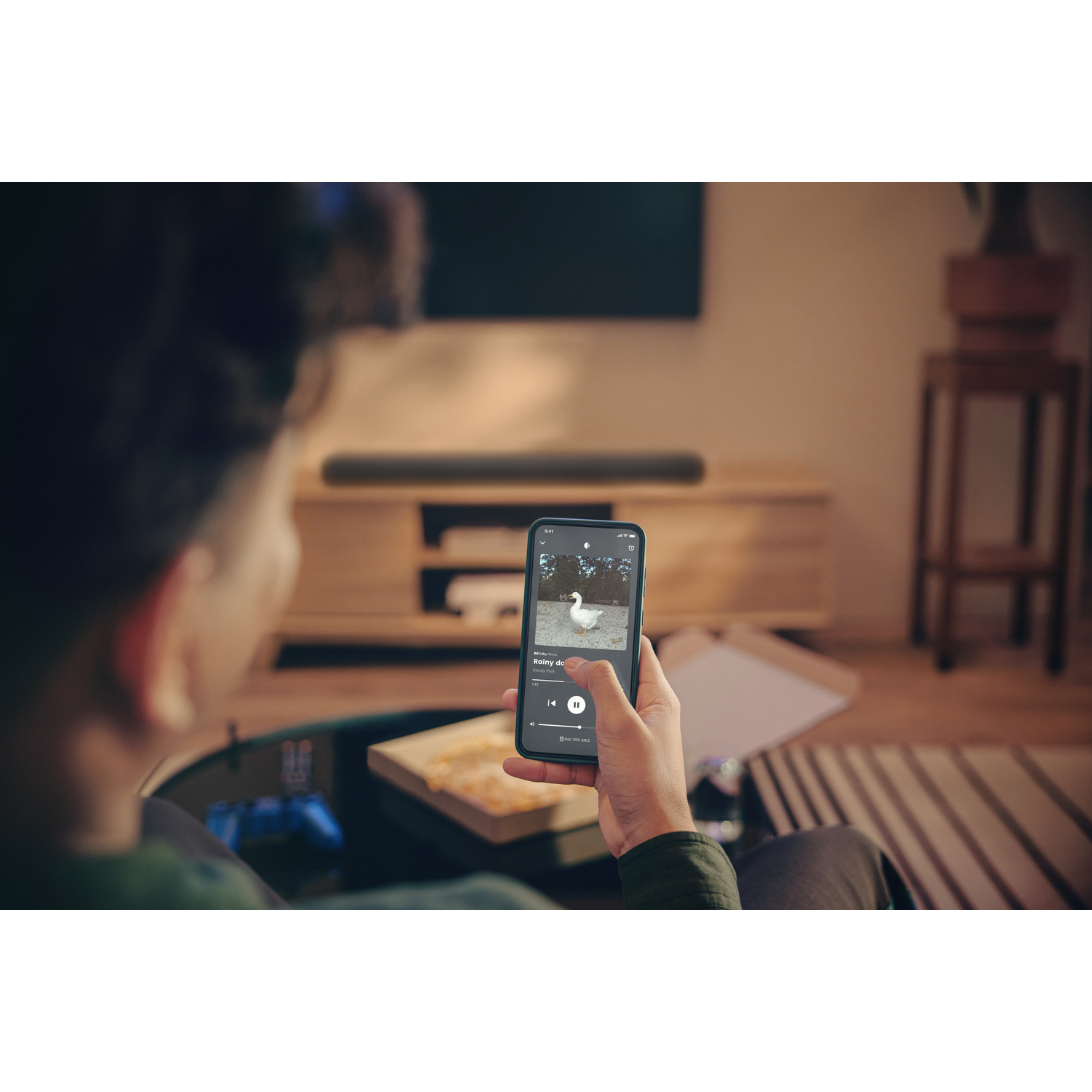 Personne tenant un téléphone jouant de la musique, image de canard à l'écran. Meuble en bois, barre de son et pièce avec tapis en arrière-plan.
