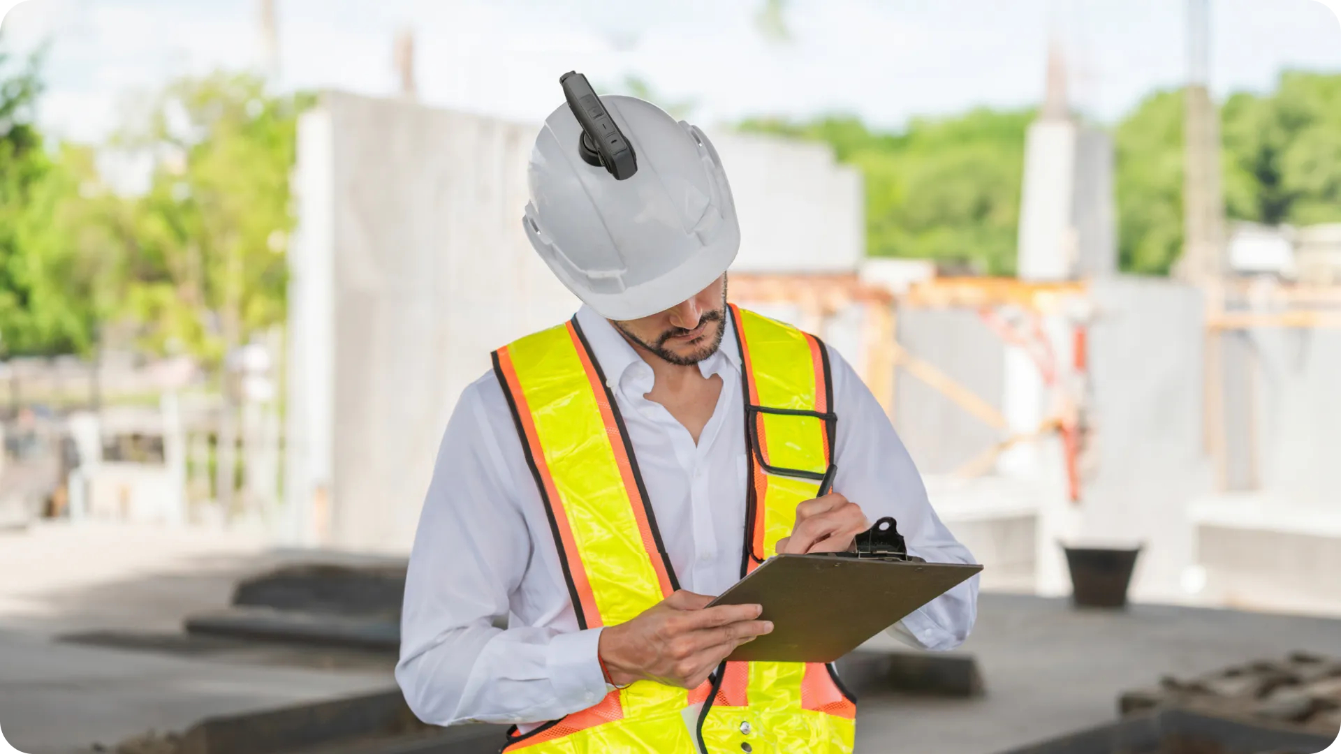 Ein Bauarbeiter mit Helm und Sicherheitsweste schreibt auf einen Klemmbrett. Im Hintergrund ist eine Baustelle zu sehen.