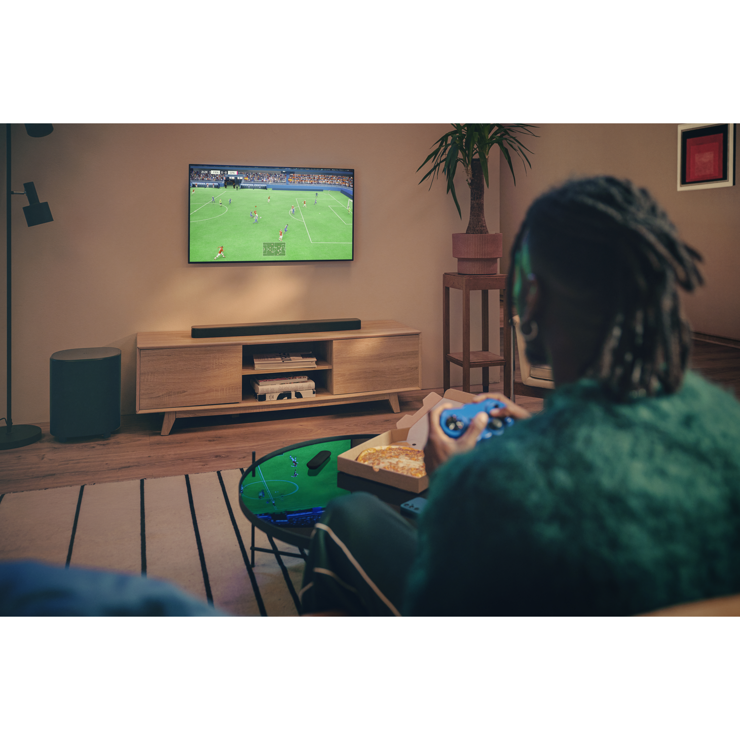 Personne jouant à un jeu vidéo avec une manette, pizza et TV avec match de foot. Ambiance salon.