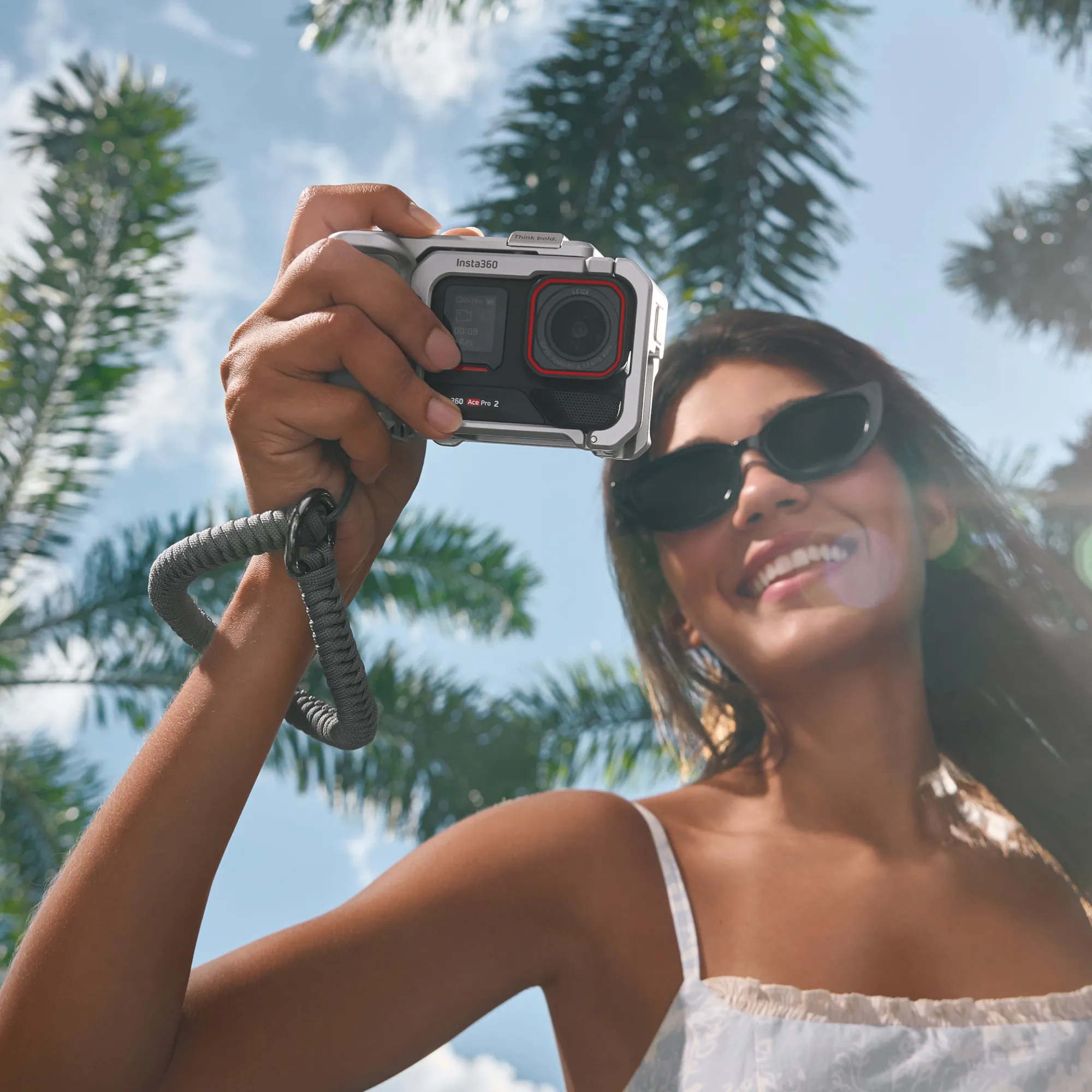 Eine Frau hält eine silberne Insta360-Kamera, Palmen, blauer Himmel, trägt Sonnenbrille. Sie lächelt in die Kamera.