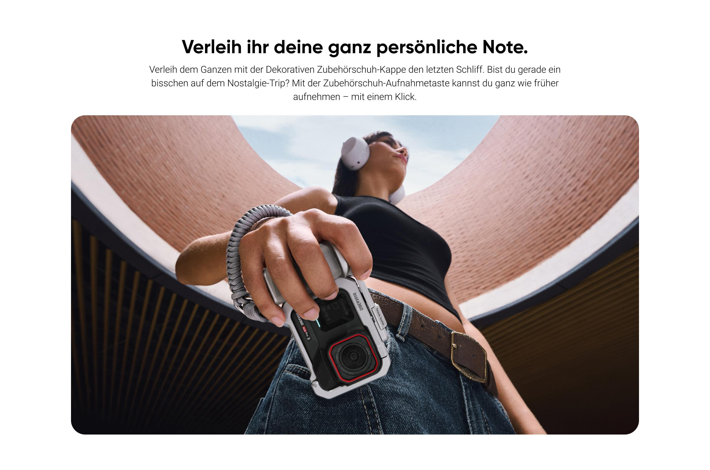 Eine Person hält eine Kamera. Sie trägt Kopfhörer und ein schwarzes Top, mit Jeans und einem braunen Gürtel. Der Hintergrund ist ein architektonisches Design.