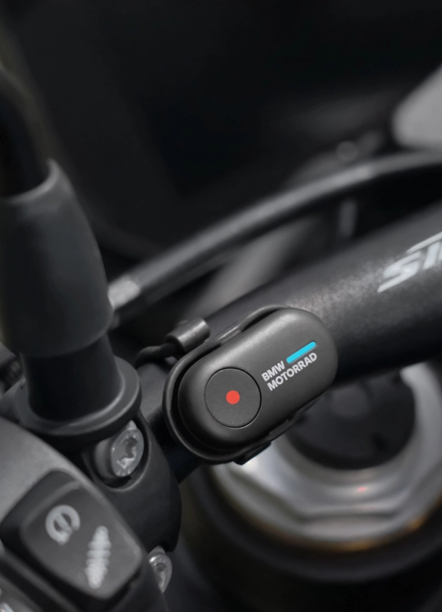 Nahaufnahme eines schwarzen Motorradlenkers mit einem BMW Motorrad Gerät. Ein rotes Licht leuchtet, Betriebsanzeige. Der Hintergrund ist unscharf.