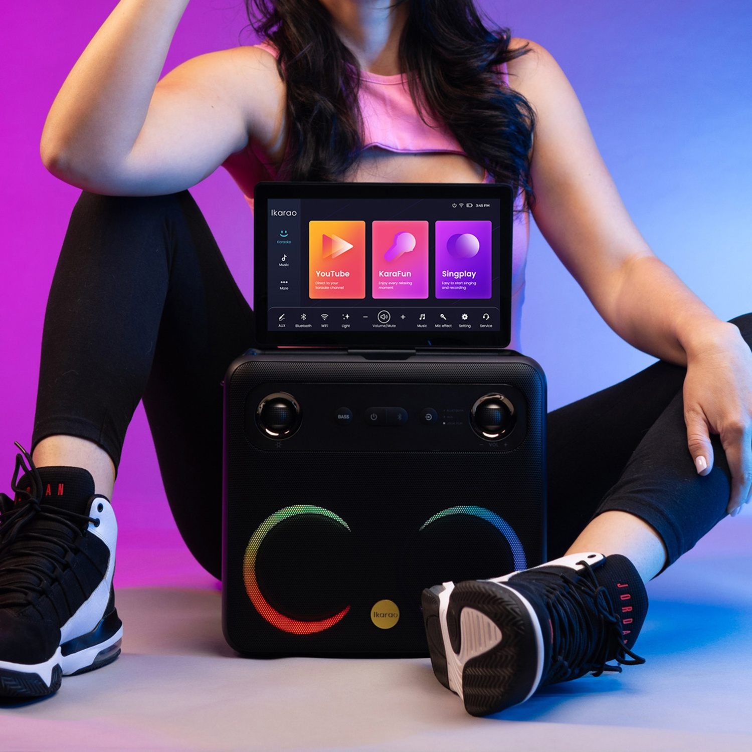 Donna seduta con macchina per karaoke. Il tablet mostra app karaoke. Altoparlante nero con luci colorate, loghi. Visibili scarpe da ginnastica e leggings neri.