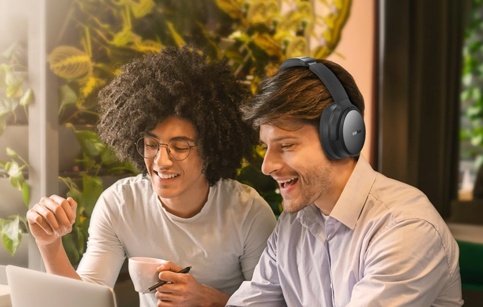 Két mosolygó férfi, egyikük fejhallgatóval, laptopot néz. Az egyikük csészét és tollat tart. Növények a háttérben.