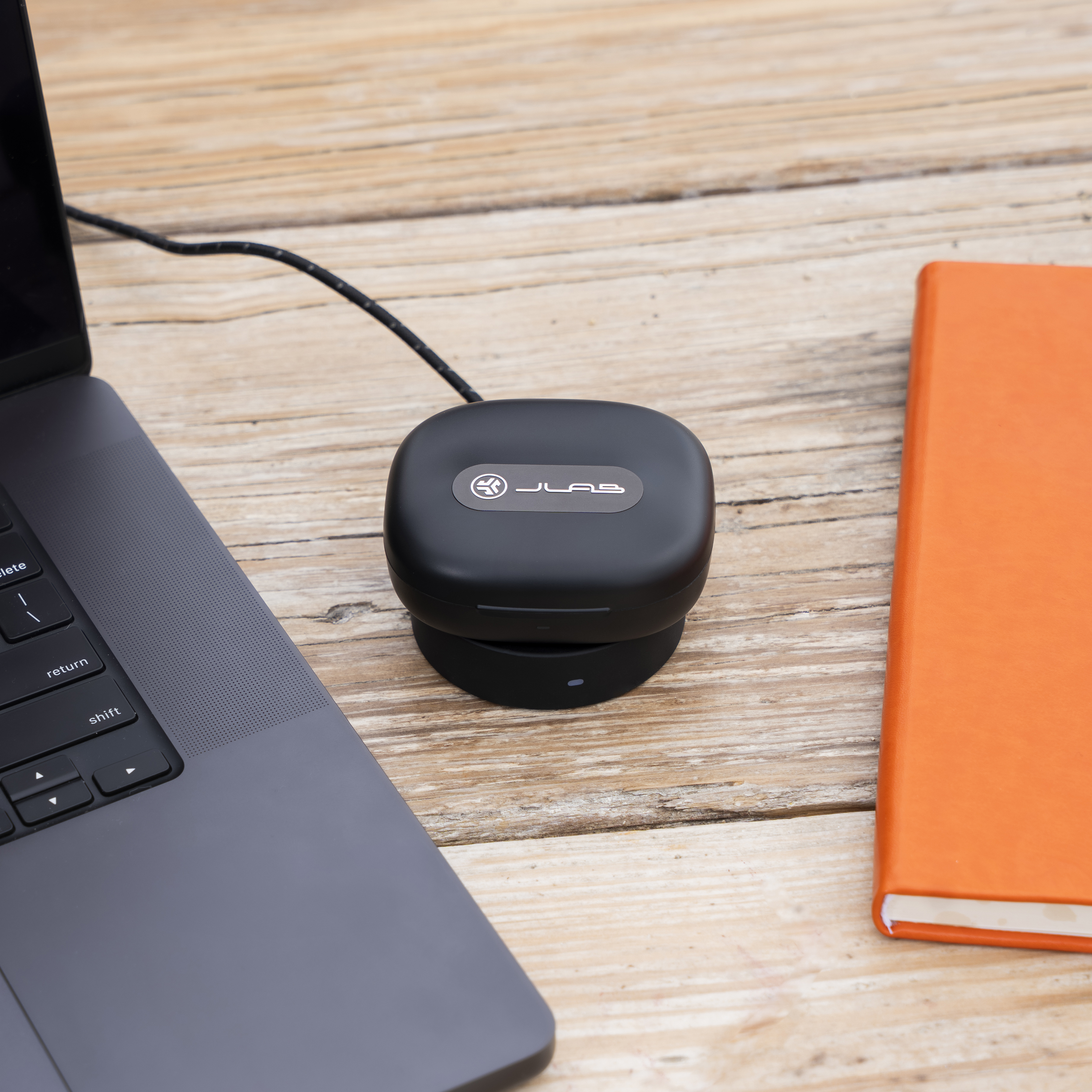 Schwarzes Ladeetui für Ohrhörer, Laptop, orangefarbenes Notizbuch auf Holzoberfläche. Kabel am Etui angeschlossen.